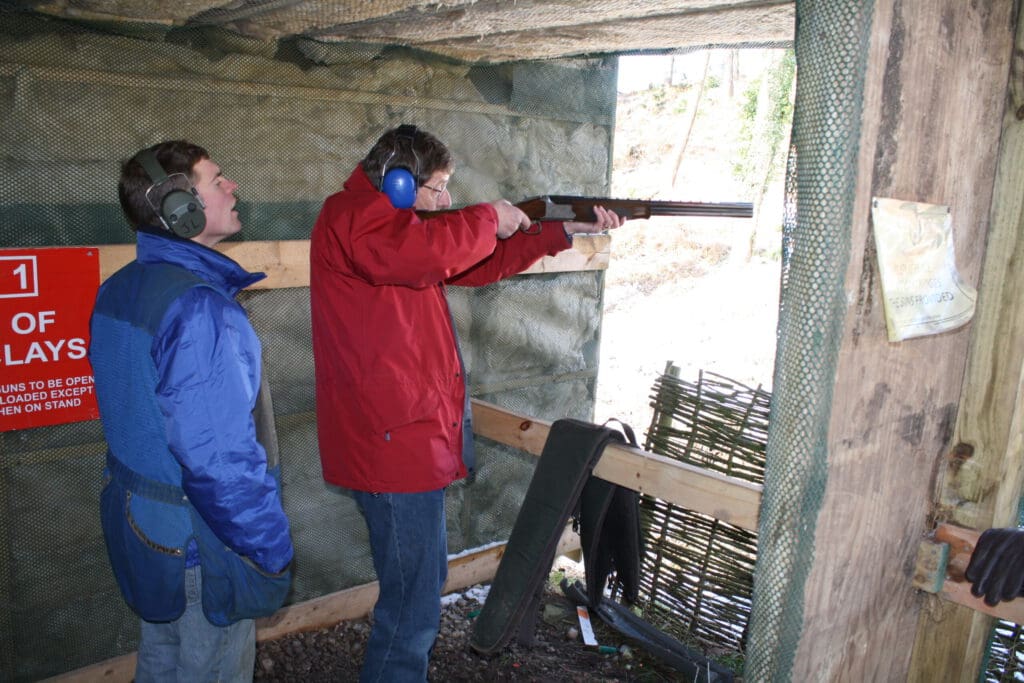 Two people wearing ear protection are at a shooting range. One person in a red jacket aims a shotgun while the other, in a blue jacket, stands beside them, supervising. A safety sign is visible on the wall.