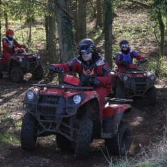 Three people wearing helmets and red riding gear drive red quad bikes along a muddy forest trail, with trees and sunlight filtering through the foliage around them.
