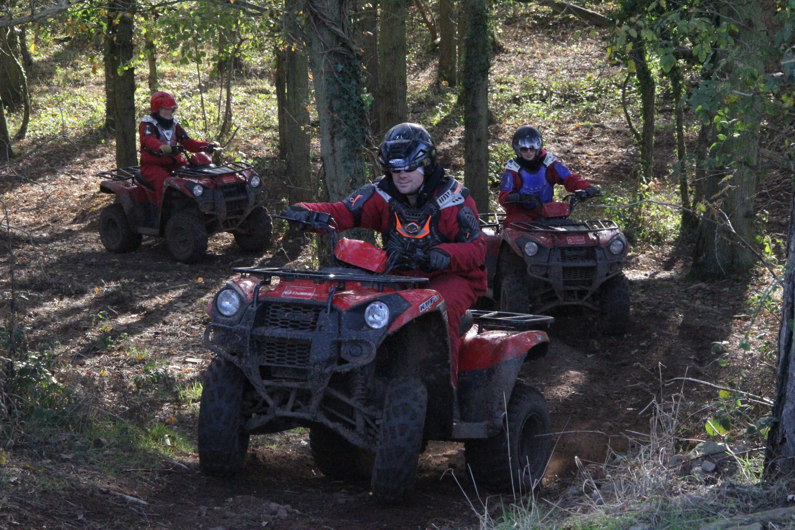 Three people wearing helmets and red riding gear drive red quad bikes along a muddy forest trail, with trees and sunlight filtering through the foliage around them.