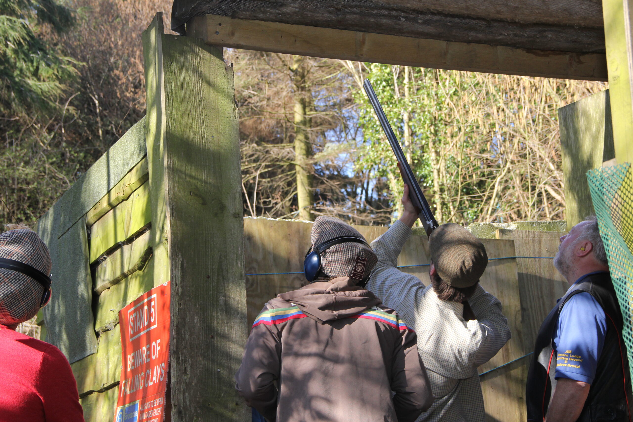 Four people stand in an outdoor wooden enclosure. One person aims a shotgun upwards, watched by others wearing protective gear. A sign on the wall reads “SHOOTING: BEWARE OF THE LAWS.” Trees are visible in the background.