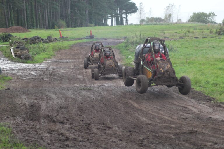 Three off-road buggies race on a muddy dirt track, with one buggy in the foreground airborne after hitting a bump, while the other two follow closely behind. Tall trees border the track on one side.
