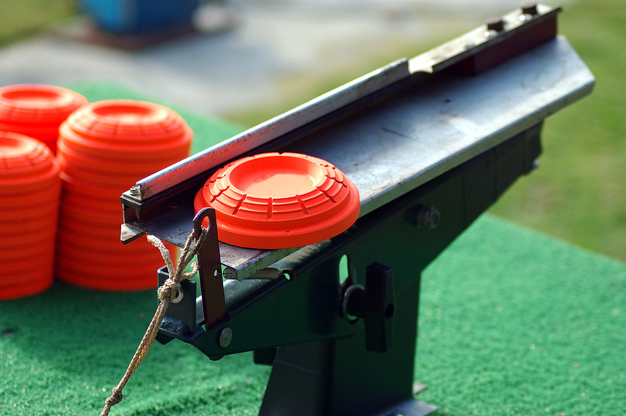 A close-up of a clay pigeon launcher with an orange clay target loaded and more orange clay targets stacked nearby, set on a green surface outdoors.