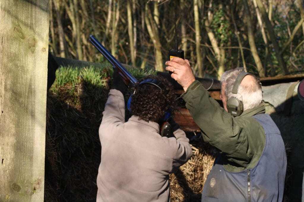 Two people at an outdoor shooting range; one is aiming a shotgun while wearing ear protection, and the other is raising a hand-held device, possibly to signal or track shots. Trees are visible in the background.
