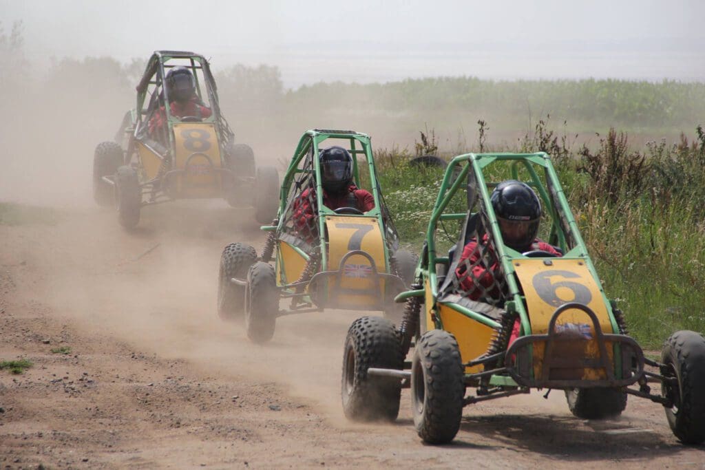Three people race off-road dune buggies numbered 6, 7, and 8 on a dirt track, kicking up dust as they speed past greenery. All drivers wear helmets and red plaid shirts.
