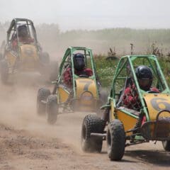 Three people race off-road dune buggies numbered 6, 7, and 8 on a dirt track, kicking up dust as they speed past greenery. All drivers wear helmets and red plaid shirts.