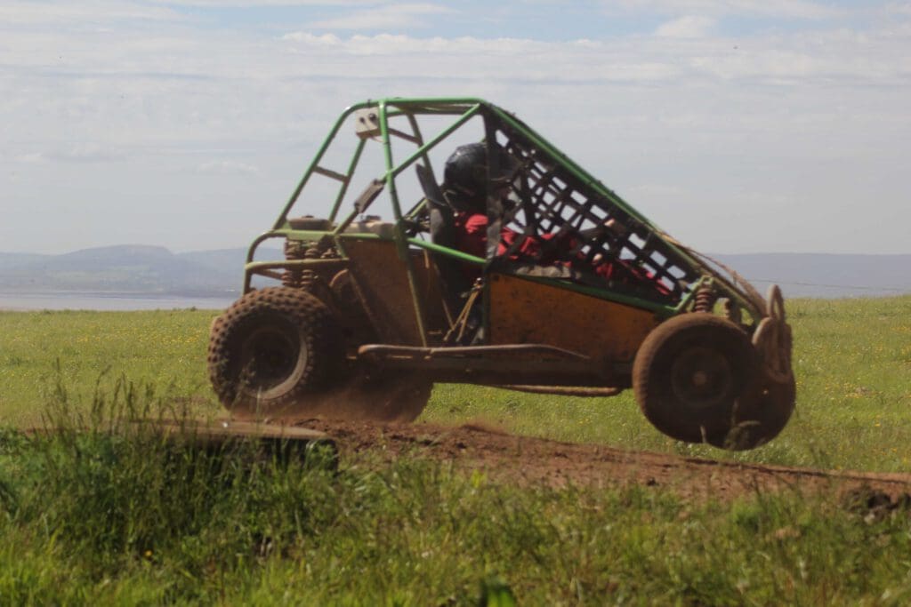 An off-road buggy with a driver wearing a helmet speeds along a dirt track, kicking up dust and catching air, with grassy fields and distant hills in the background.