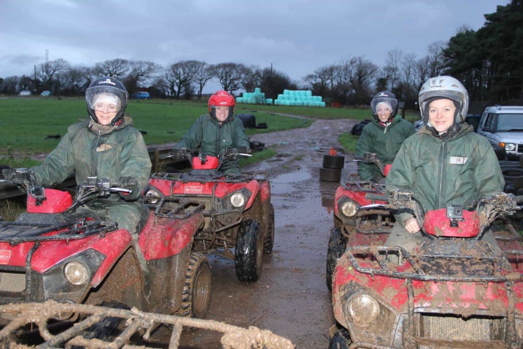 Four people in green jackets and helmets sit on muddy red quad bikes on a dirt track, smiling at the camera. The background features grassy fields, stacks of hay bales, and trees under a cloudy sky.