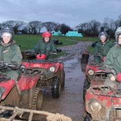 Four people in green jackets and helmets sit on muddy red quad bikes on a dirt track, smiling at the camera. The background features grassy fields, stacks of hay bales, and trees under a cloudy sky.