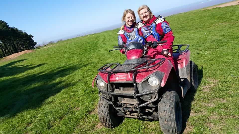 Two women in red overalls smiling while sitting on a red quad bike on a grassy field under a clear blue sky, with trees visible in the background.