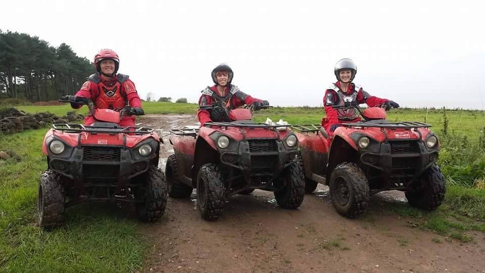 Three people wearing helmets and red jackets sit on muddy red quad bikes, smiling and posing on a dirt path in a grassy outdoor area with trees in the background.