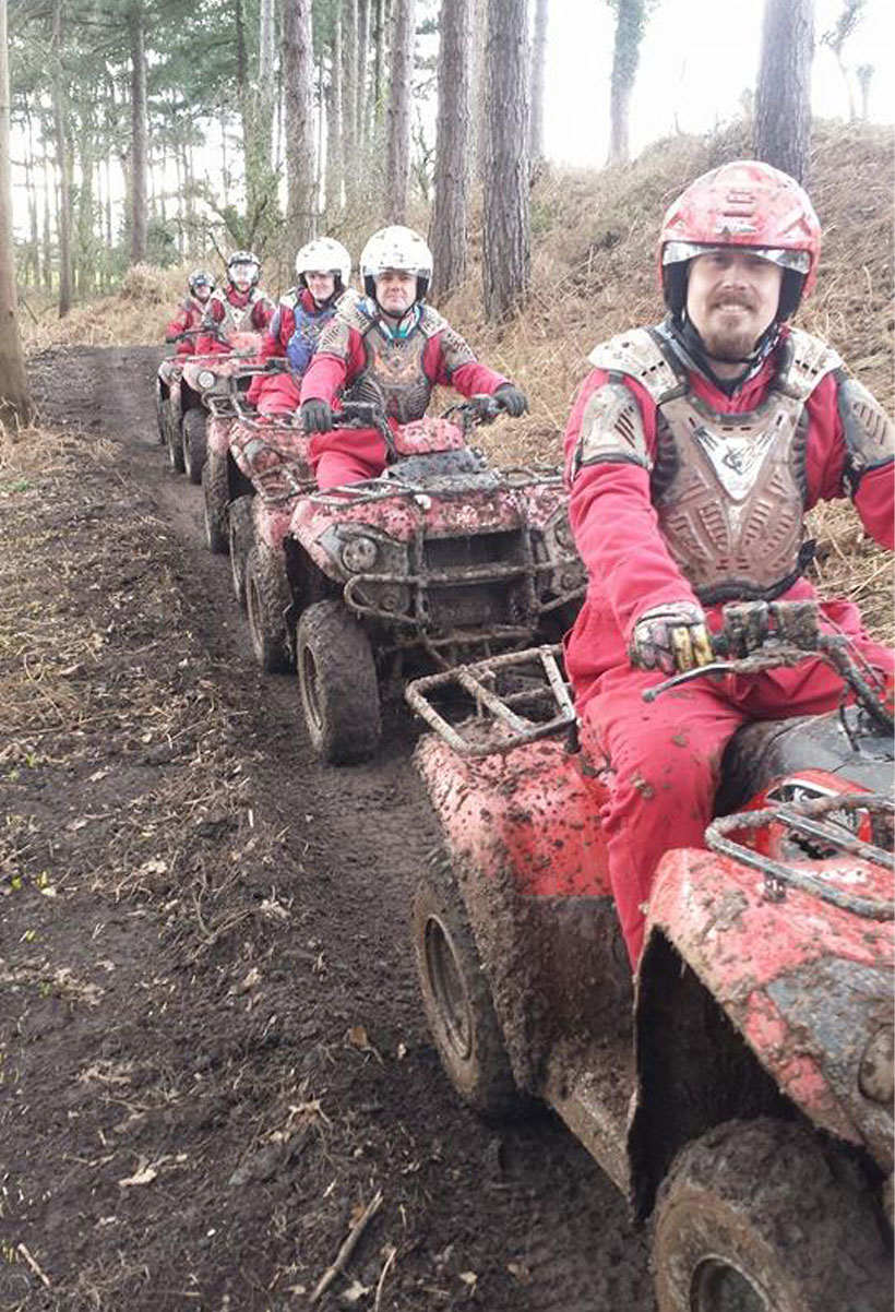 Five people in red protective gear and helmets ride muddy quad bikes in a single file on a dirt path through a wooded area. The riders appear to be participating in an outdoor adventure or off-road activity.