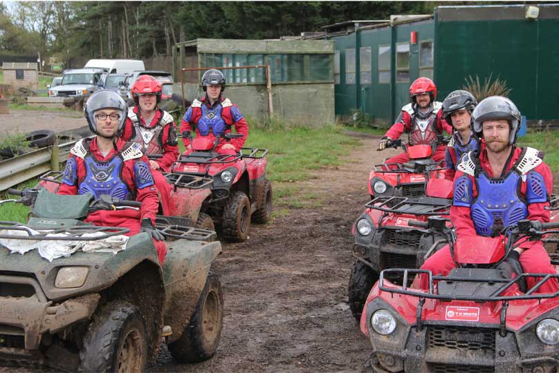 A group of six people wearing helmets and protective gear sit on muddy quad bikes, ready to ride on a dirt track with green buildings and trees in the background.