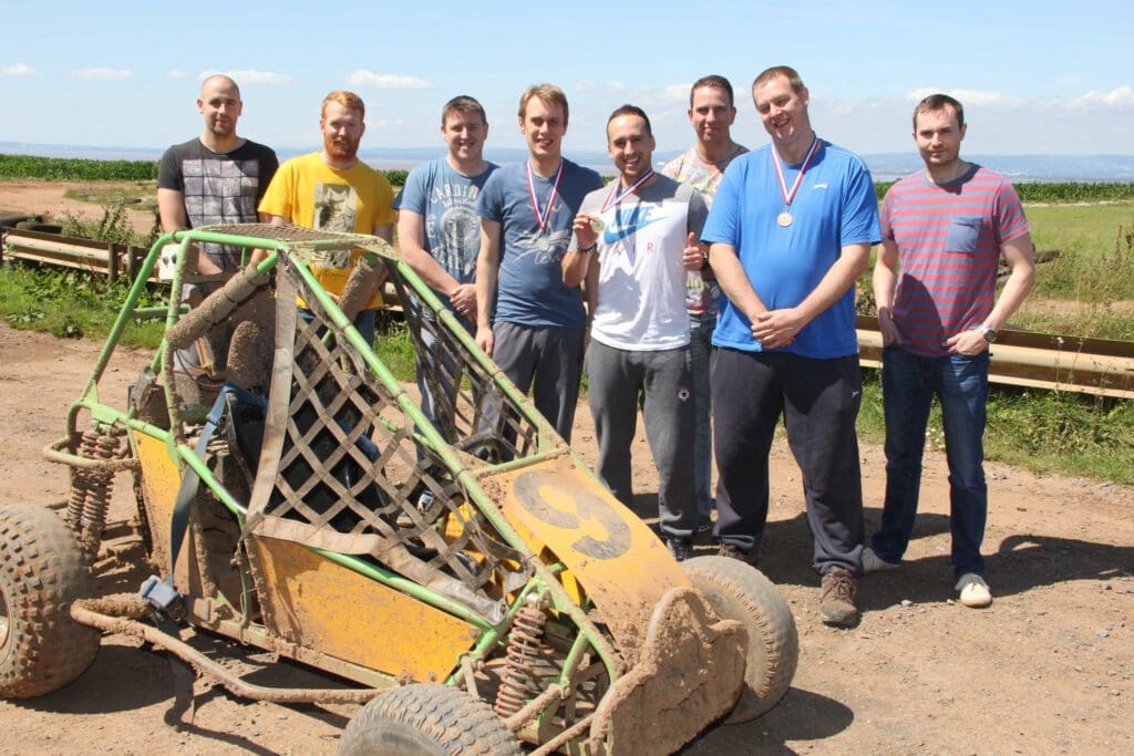 Eight men stand outdoors behind a muddy, yellow and green off-road buggy. Some are wearing medals and smiling, with fields and a blue sky in the background.