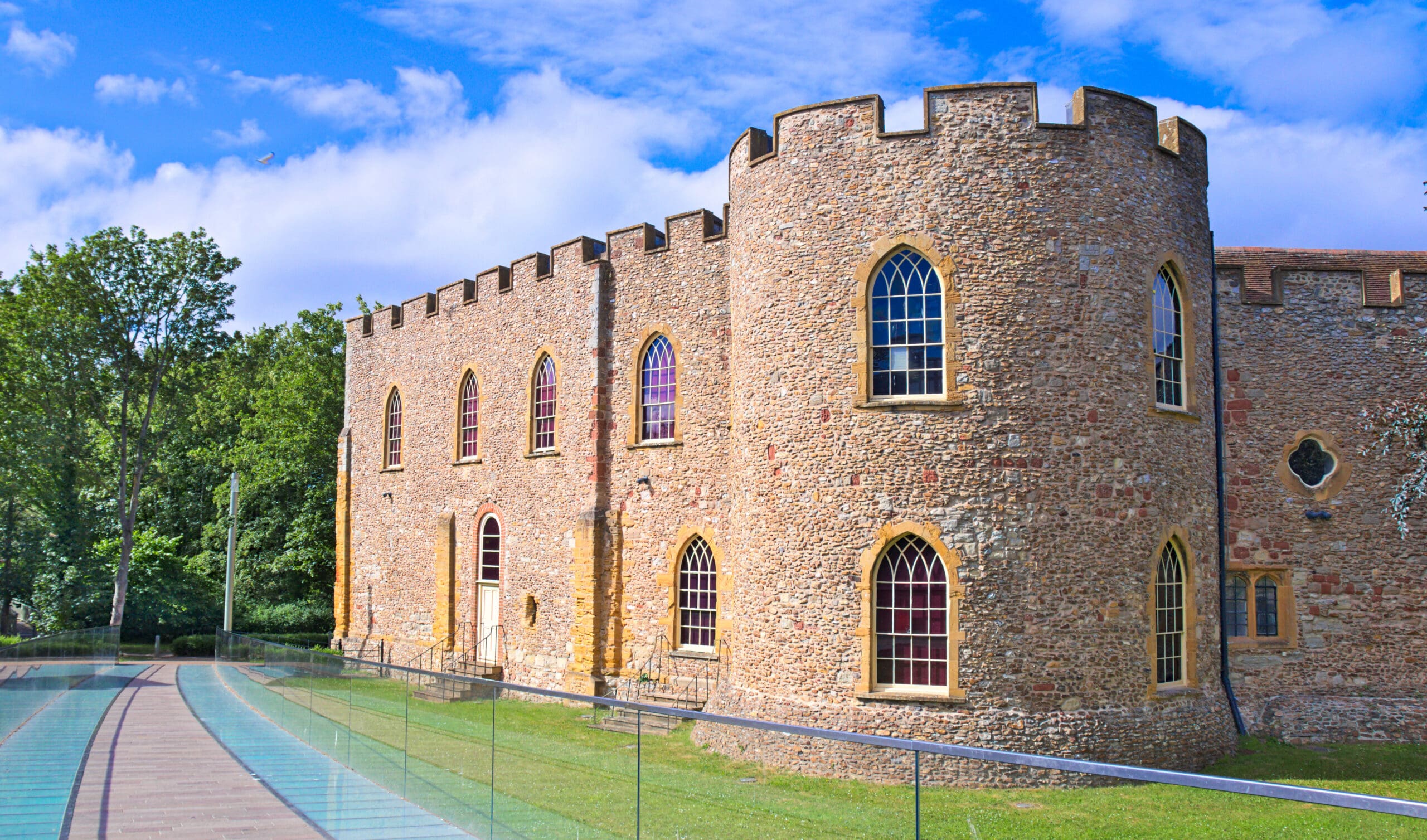 A historic stone castle with arched windows and a crenellated tower stands next to a modern glass walkway, surrounded by grass and trees under a blue sky with scattered clouds.