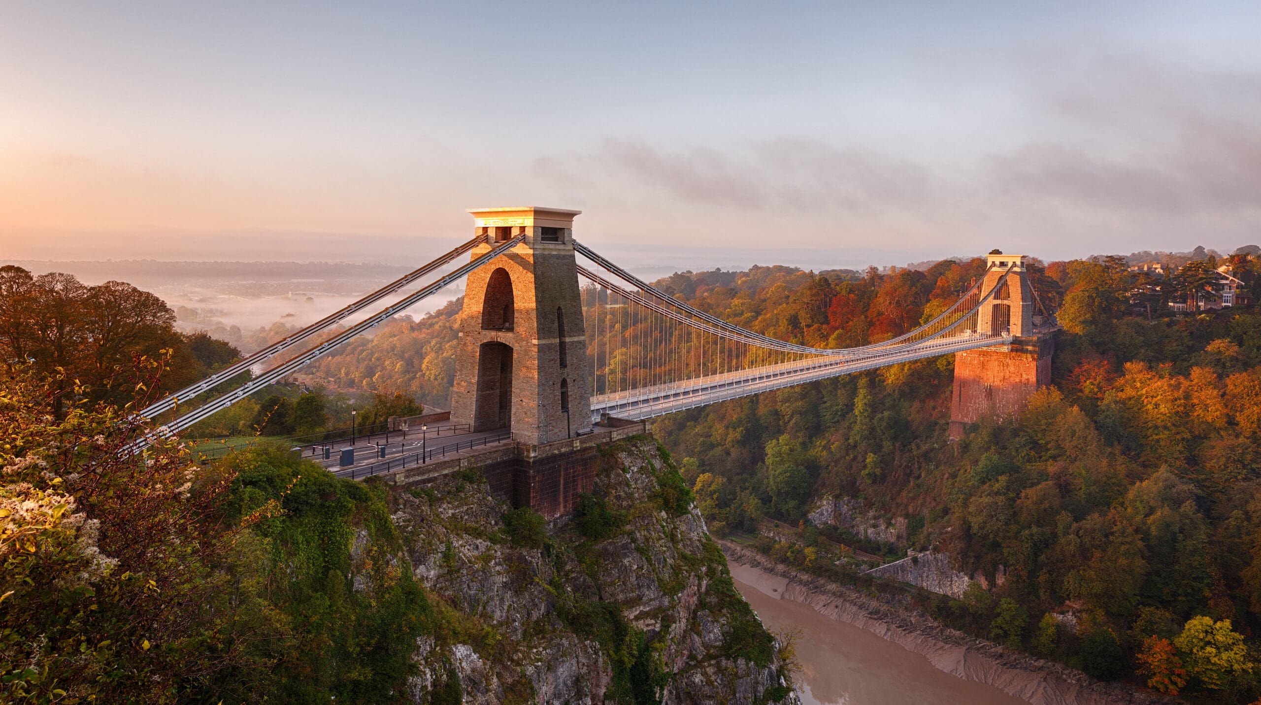 The Clifton Suspension Bridge spans a deep gorge, surrounded by autumnal trees, under a soft sunrise sky. The stone towers and steel cables are reflected in the calm river below.