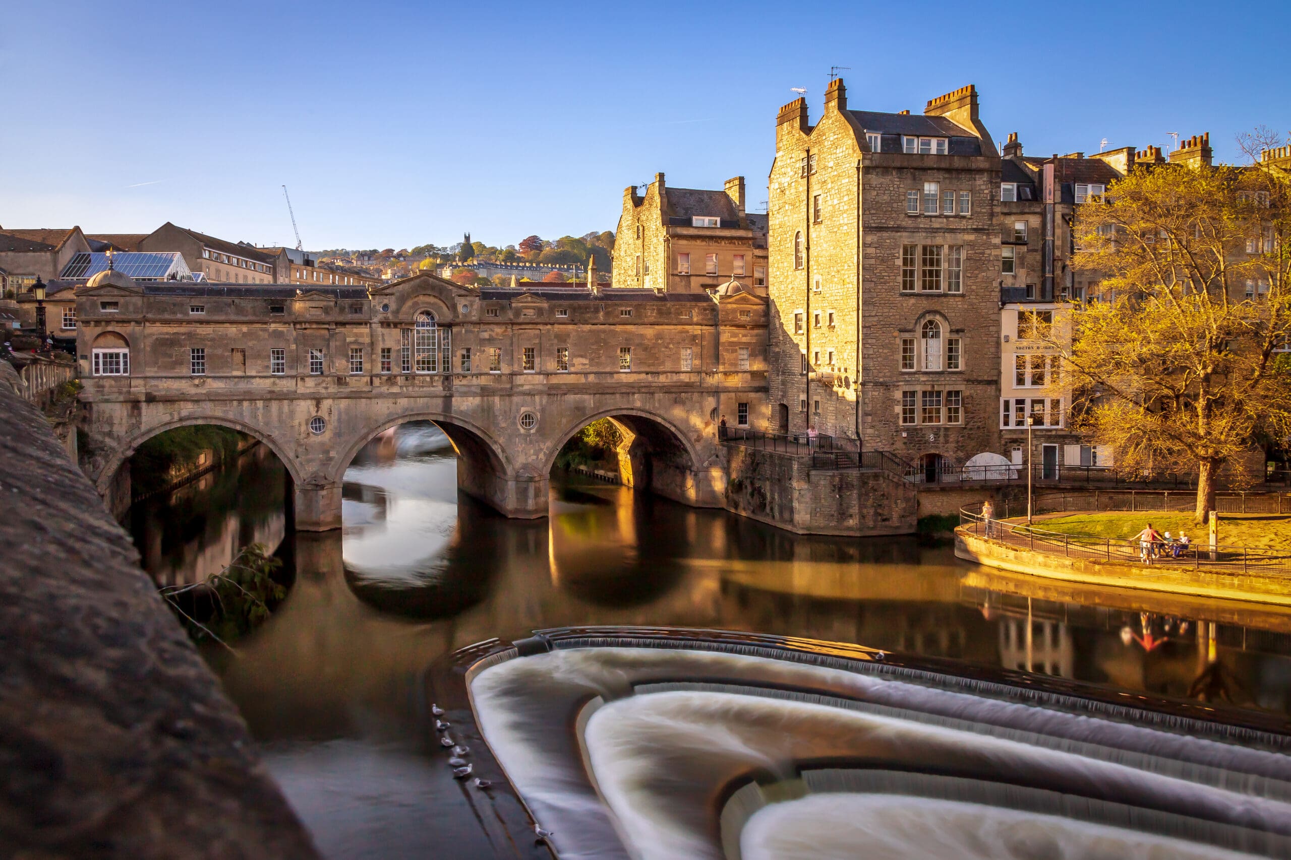 Historic stone bridge with shops, known as Pulteney Bridge, spans a calm river with a curved weir. Old buildings line the water under a clear blue sky, and sunlight bathes the scene in a warm glow.
