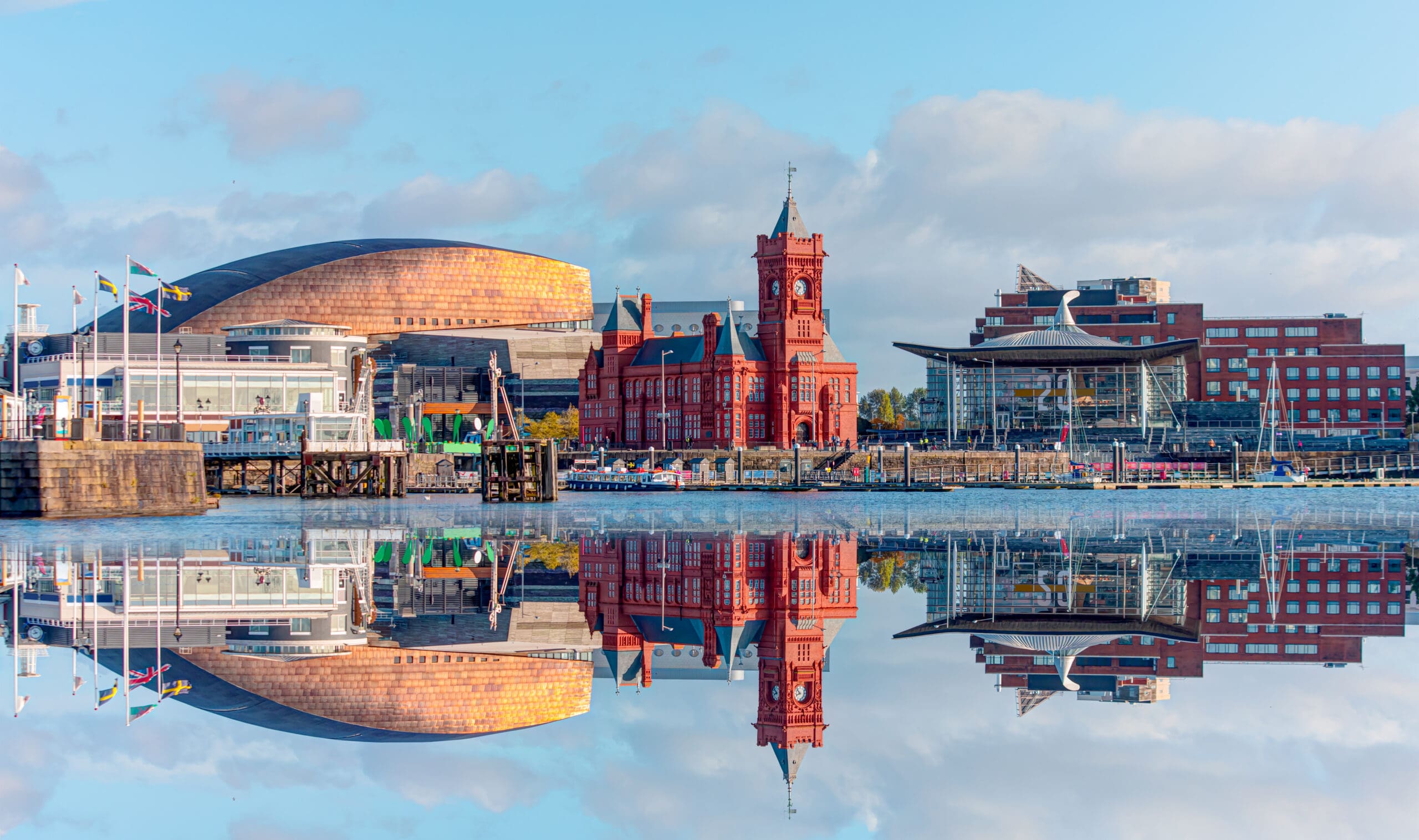 A view of Cardiff Bay with modern and historic buildings reflected in the calm water, including the Wales Millennium Centre and the red Pierhead Building, under a blue sky with scattered clouds.