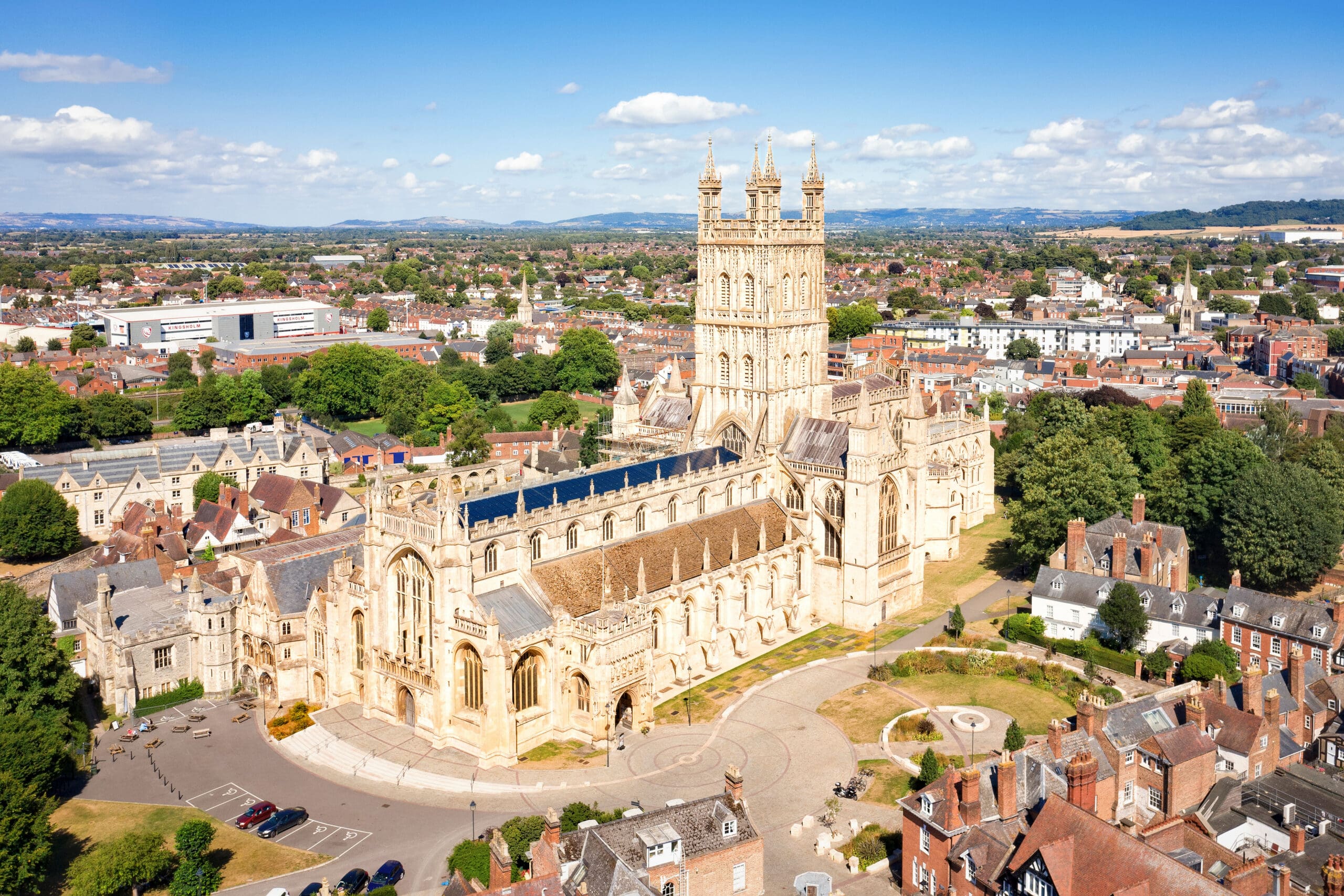 Aerial view of Gloucester Cathedral, a large historic stone building with towers and intricate architecture, surrounded by trees and city buildings under a bright blue sky.