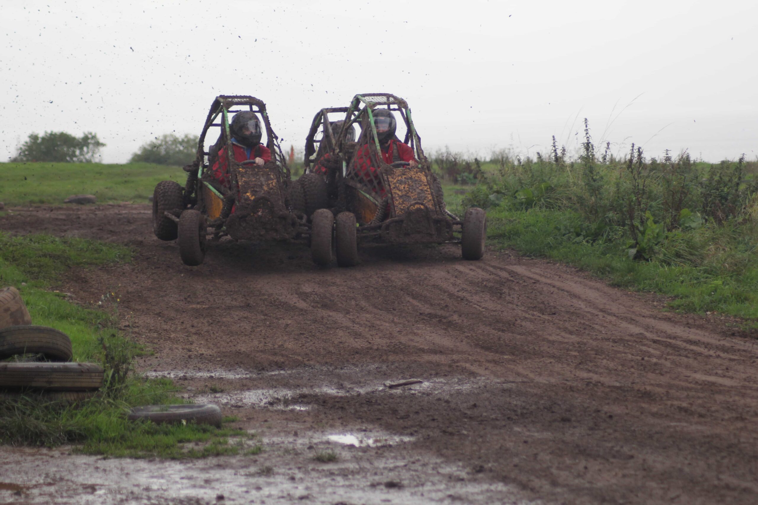 Two off-road buggies, covered in mud, race side by side on a dirt track with green grass and bushes on either side. Both drivers wear helmets and red outfits as mud flies up from the tires.