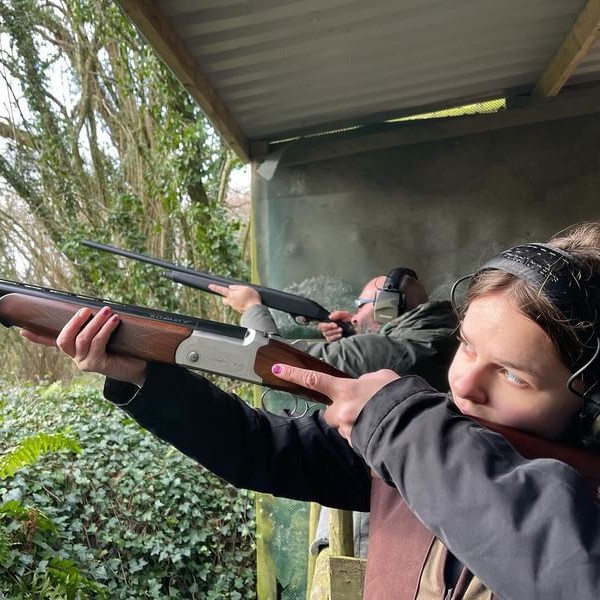 Two people wearing ear protection aim shotguns at an outdoor shooting range. The girl in the foreground focuses intently, surrounded by greenery and a covered structure above.