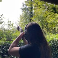 A person with long hair aims a shotgun toward the sky in a lush, green outdoor setting surrounded by trees and foliage.