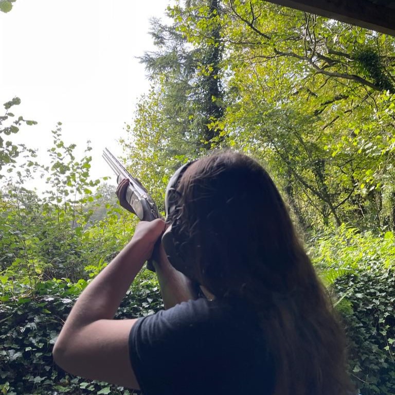 A person with long hair aims a shotgun toward the sky in a lush, green outdoor setting surrounded by trees and foliage.