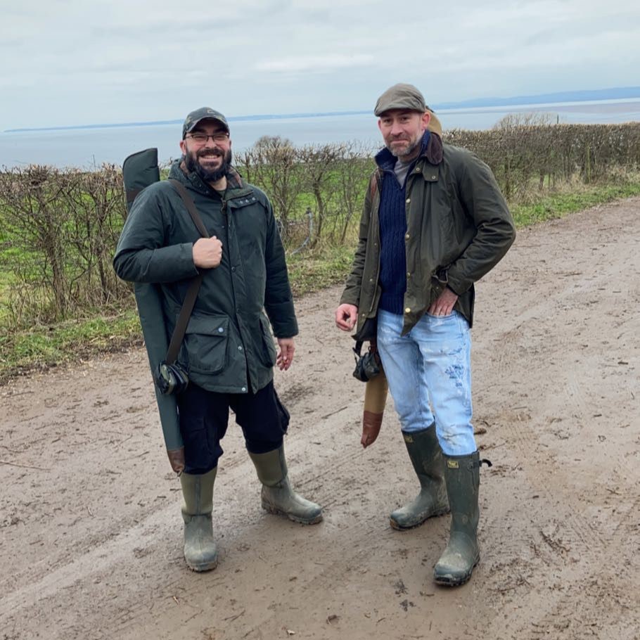 Two men wearing jackets, rubber boots, and hats stand on a muddy rural path. One man holds a green case over his shoulder. There are bushes and a glimpse of water in the background under a cloudy sky.