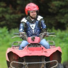 A man wearing a red helmet and blue protective gear rides a muddy red ATV outdoors, with green foliage blurred in the background.