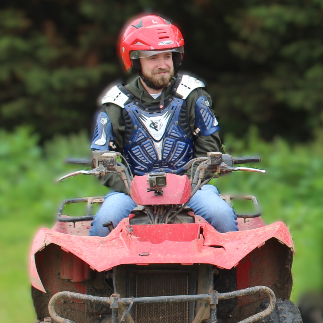 A man wearing a red helmet and blue protective gear rides a muddy red ATV outdoors, with green foliage blurred in the background.