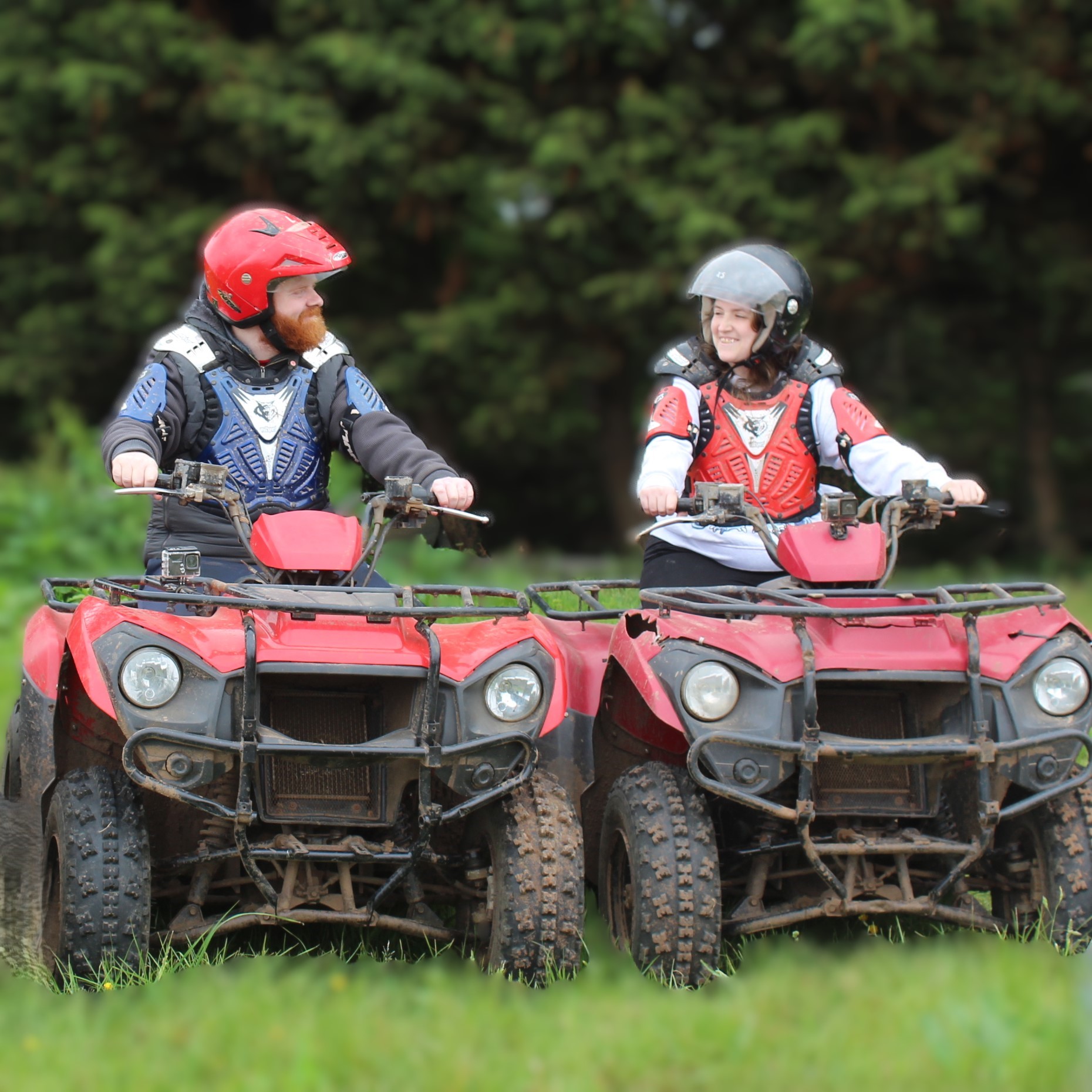 Two people wearing helmets, protective gear, and casual clothes are sitting on muddy red ATVs outdoors, facing each other and smiling, with green trees and grass in the background.