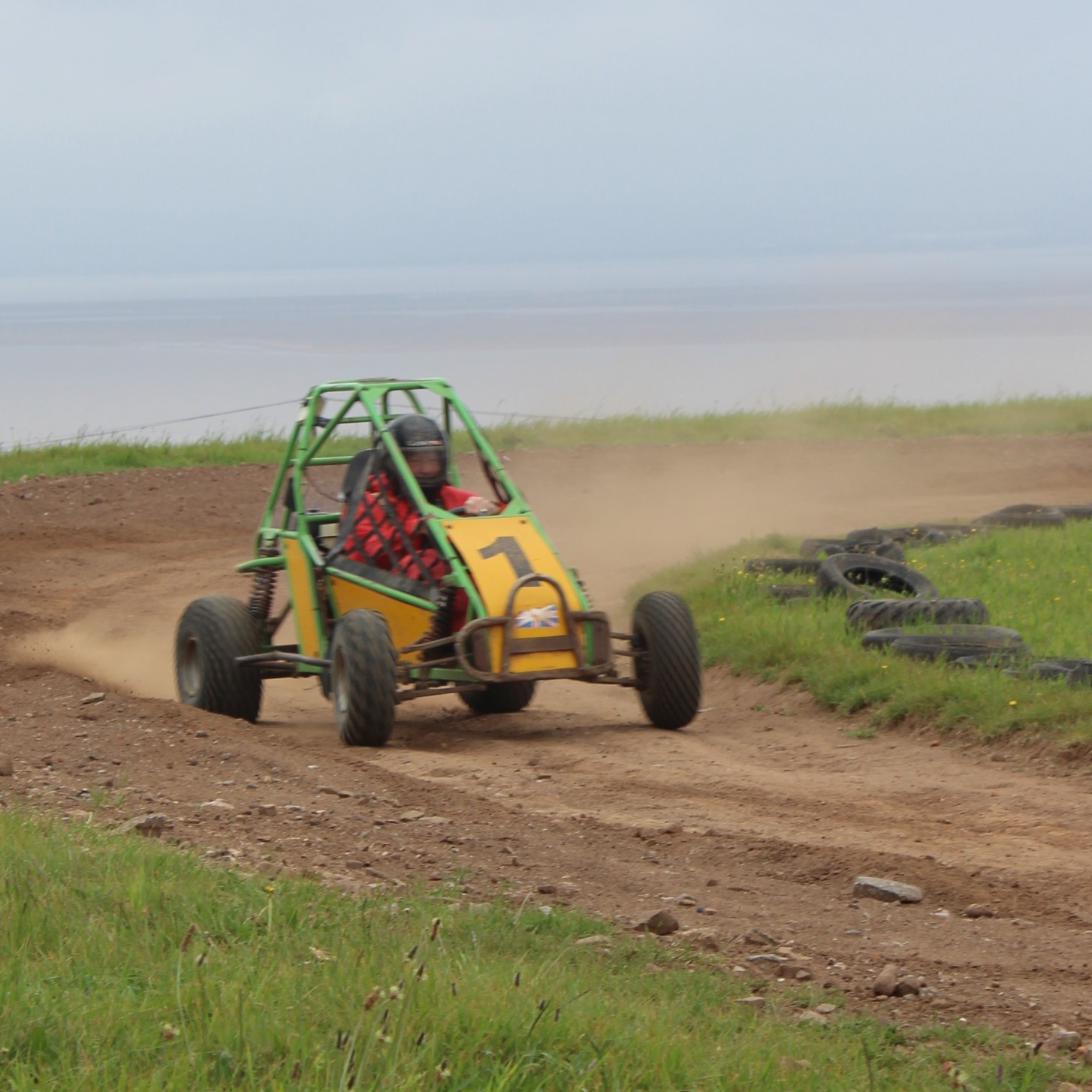 A person wearing a helmet and red jacket drives a yellow and green off-road buggy on a dirt track, kicking up dust. Old tires line the track, with grass and open sky in the background.