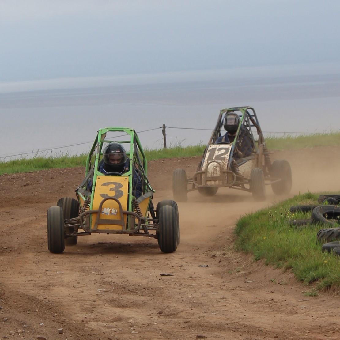Two people drive off-road buggies with roll cages, racing on a dirt track near grassy areas and water in the background. The buggy in front is yellow with the number 3; the second one is white with the number 12.