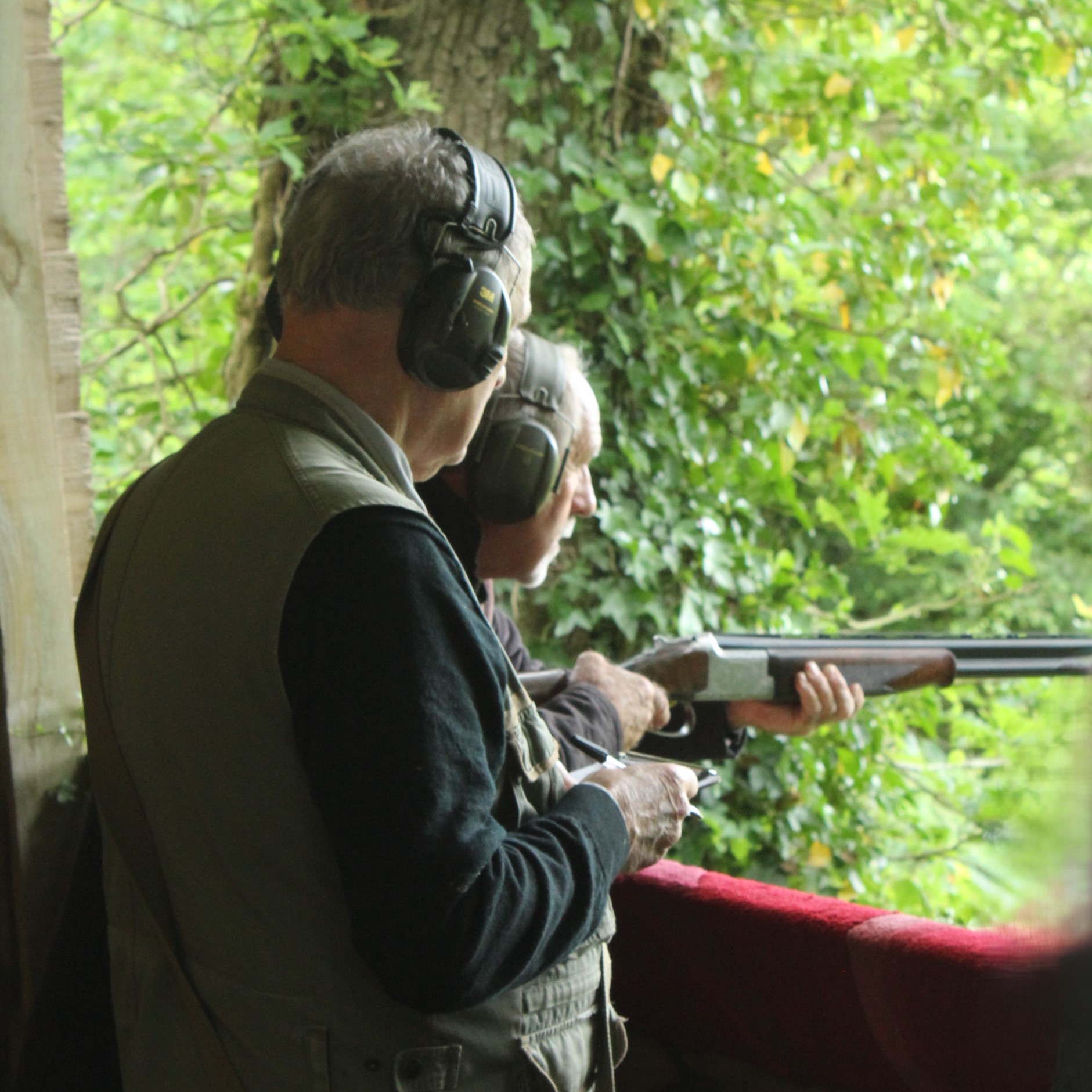 Two older men wearing ear protection stand outdoors by a wooden railing, one aiming a shotgun into a lush, green forest while the other looks on, possibly instructing or supervising.