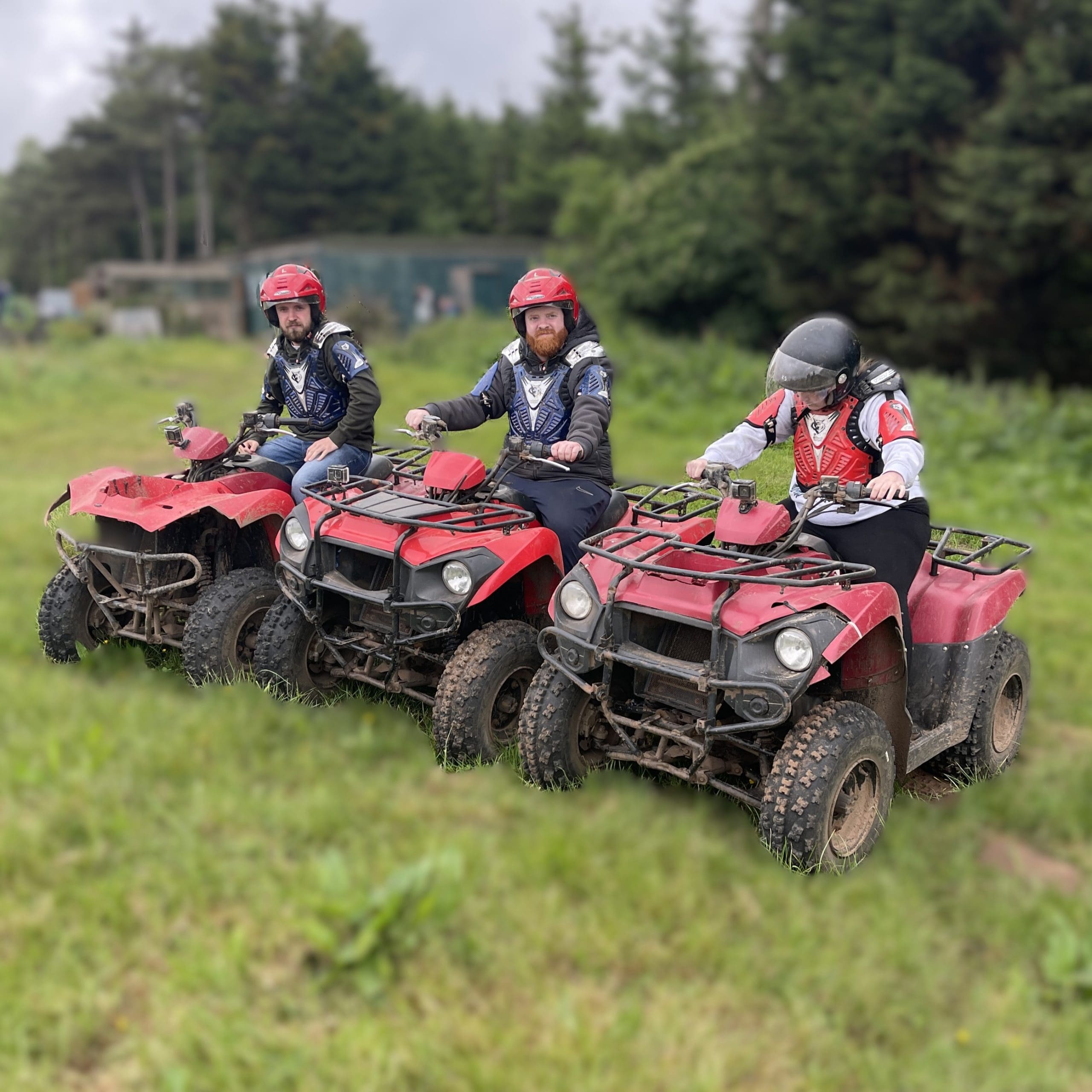 Three people in helmets and protective vests sit on red quad bikes in a grassy outdoor area with trees in the background. The ground and the vehicles are muddy.