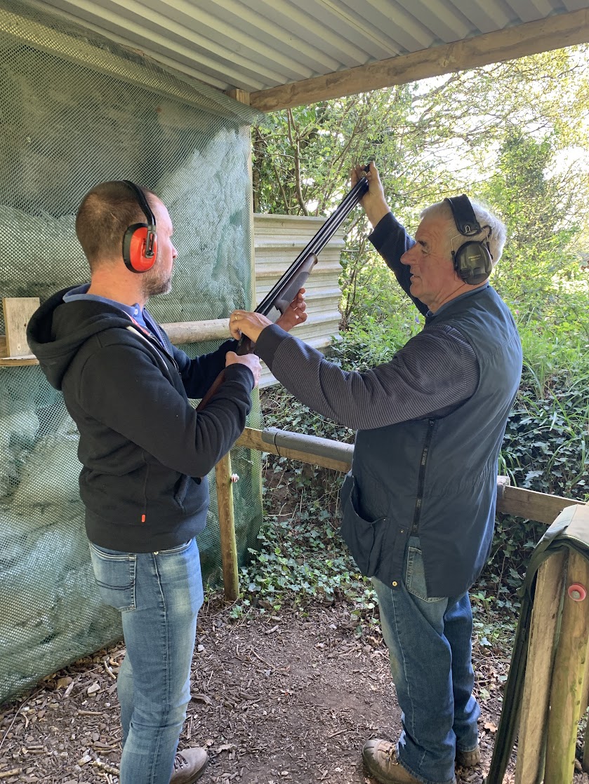 Two men wearing ear protection stand in an outdoor firing range. One man instructs the other on holding a shotgun, surrounded by greenery and standing under a metal roof.
