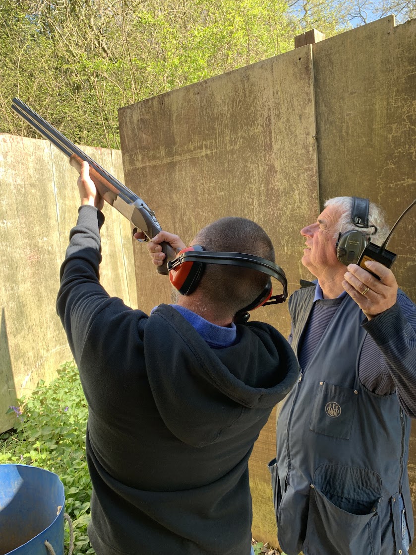 A person wearing ear protection aims a shotgun upwards, assisted by an instructor who holds a device and watches. They are outdoors, next to a wooden fence with greenery in the background.
