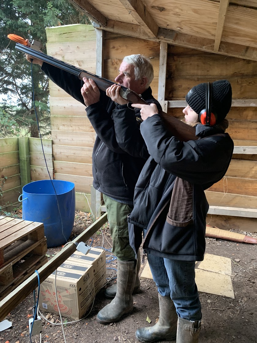 Two people stand in a wooden shed holding a shotgun. The older man is aiming while the younger, wearing ear protection, assists. Both wear jackets and boots. Nearby are a blue barrel, a cardboard box, and some wooden planks.