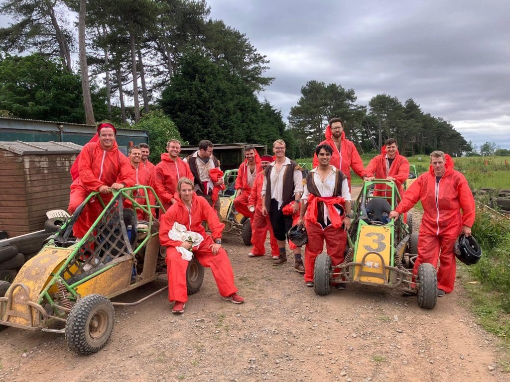 A group of people in red coveralls pose outdoors by two off-road buggies, holding helmets and smiling, with trees and cloudy skies in the background.