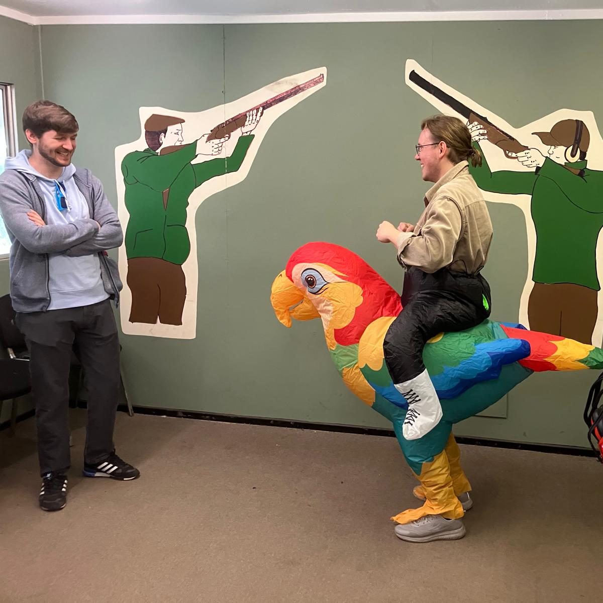 Two people are indoors; one stands smiling with arms crossed, while the other wears an inflatable parrot costume. Behind them, there are wall illustrations of hunters aiming rifles.