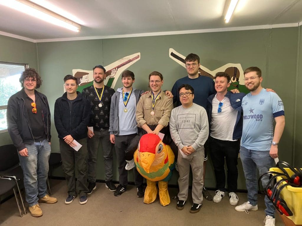 Nine people stand together indoors, some wearing medals. A large colorful bird plush toy is in front of them. The group is smiling, and two cricket bats are mounted on the green wall behind them.
