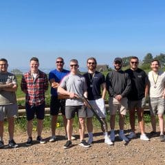 A group of ten men stand outdoors in a line on a gravel path, some smiling, some wearing sunglasses. Two men have medals, and one in the center holds a shotgun. Green grass and blue sky are in the background.