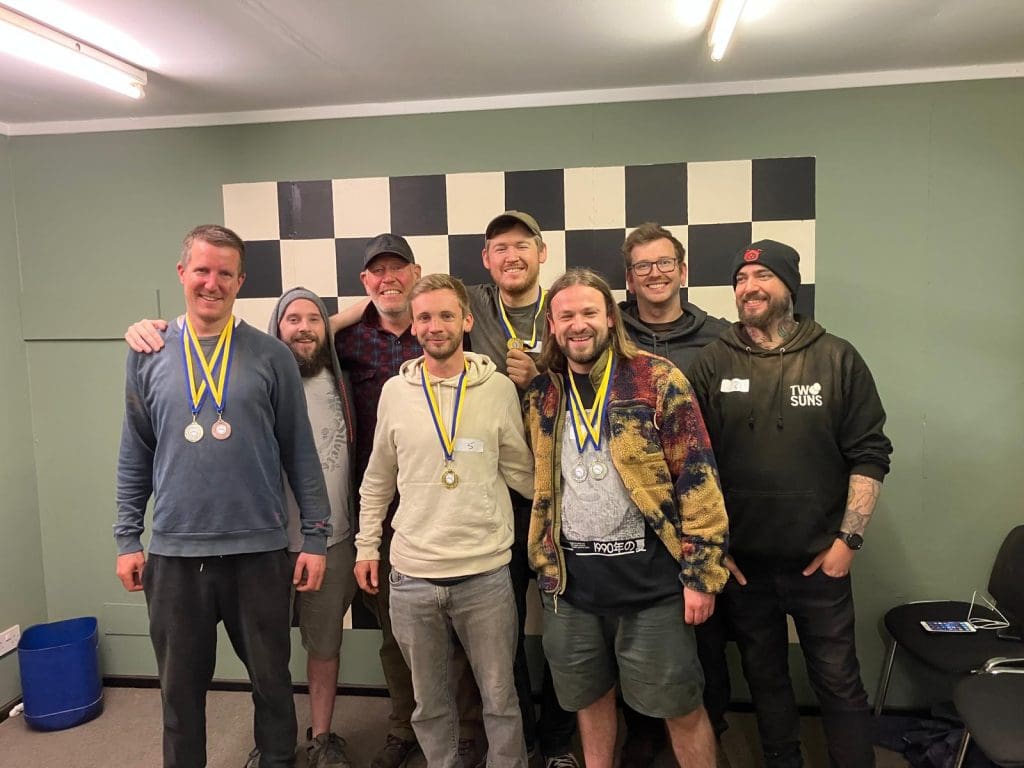 A group of seven smiling men pose indoors in front of a large black and white checkered board, with several wearing medals around their necks. They appear to be celebrating a team achievement or competition.