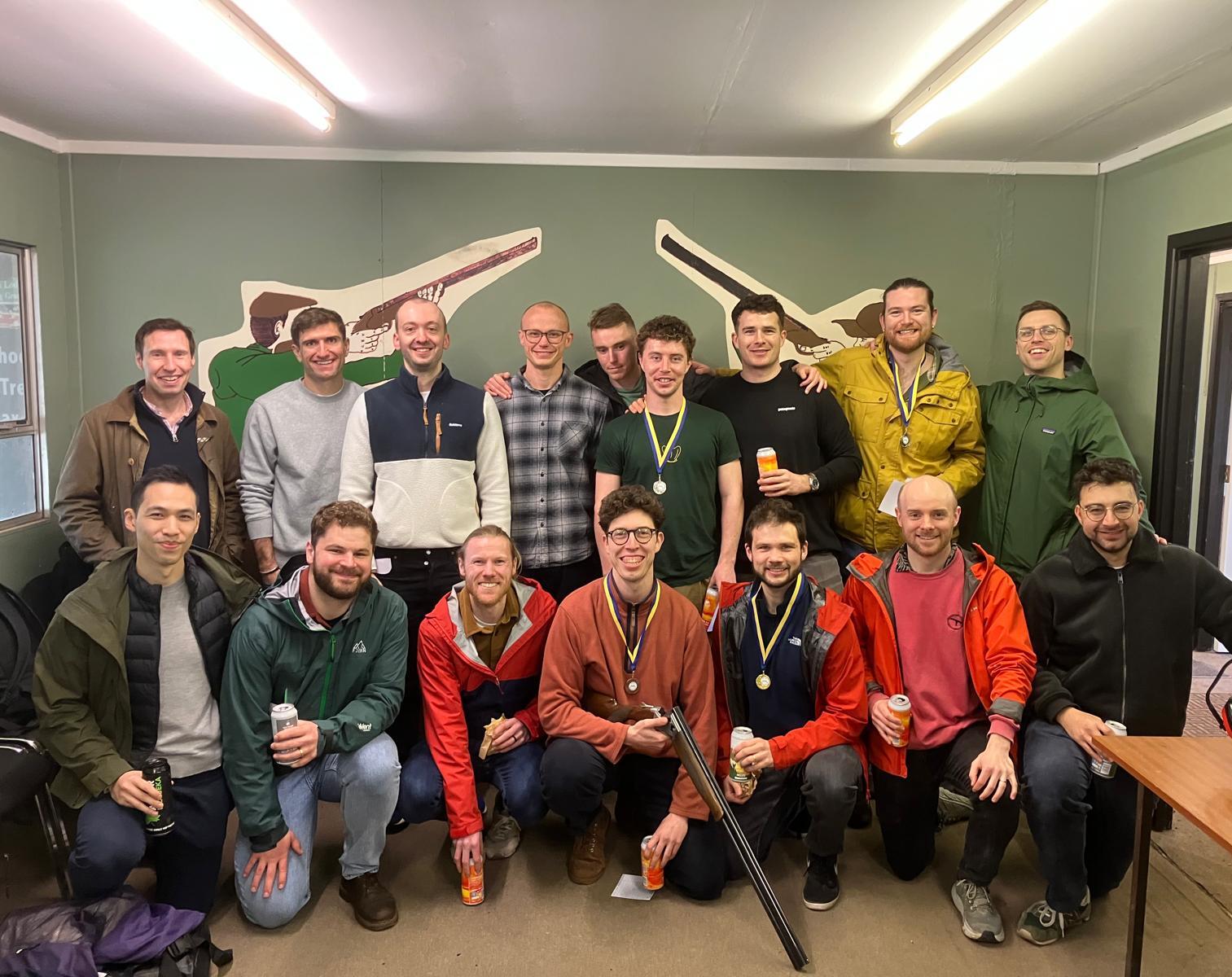 A group of sixteen smiling people, some wearing medals, pose indoors in front of a green wall with two painted rifles. Several hold drinks, and one person holds a shotgun. Most wear jackets or sweaters.