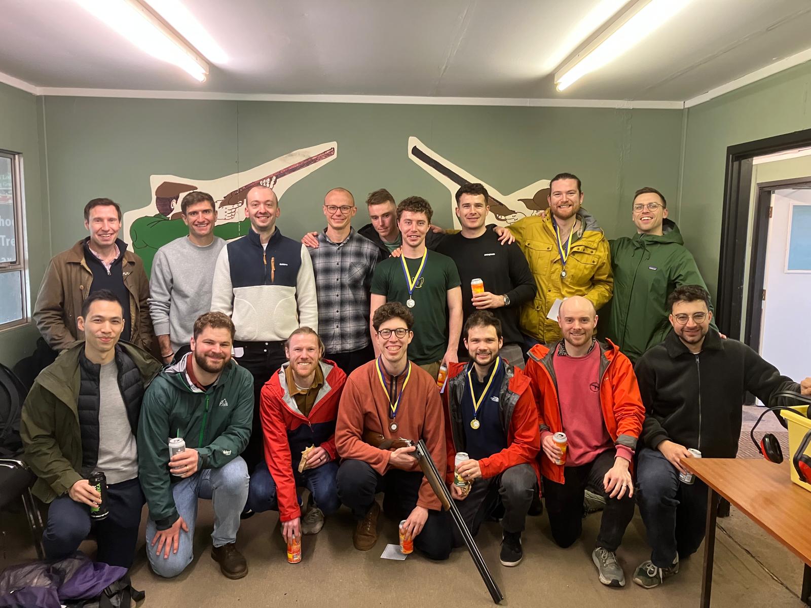 A group of sixteen people, mostly men, pose indoors, some wearing medals and holding drinks. Two large rifle silhouettes are mounted on the green wall behind them. Several people are dressed in outdoor jackets.