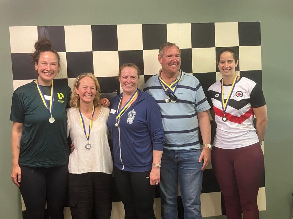 Five people wearing medals stand smiling in front of a large black and white checkered wall. Four women and one man are dressed casually, posing together for a group photo.