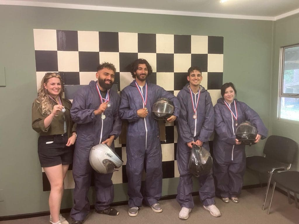 Five people stand in front of a large black-and-white checkered wall. Four are wearing blue racing suits and medals, holding helmets, while one person in casual clothes stands smiling on the left.