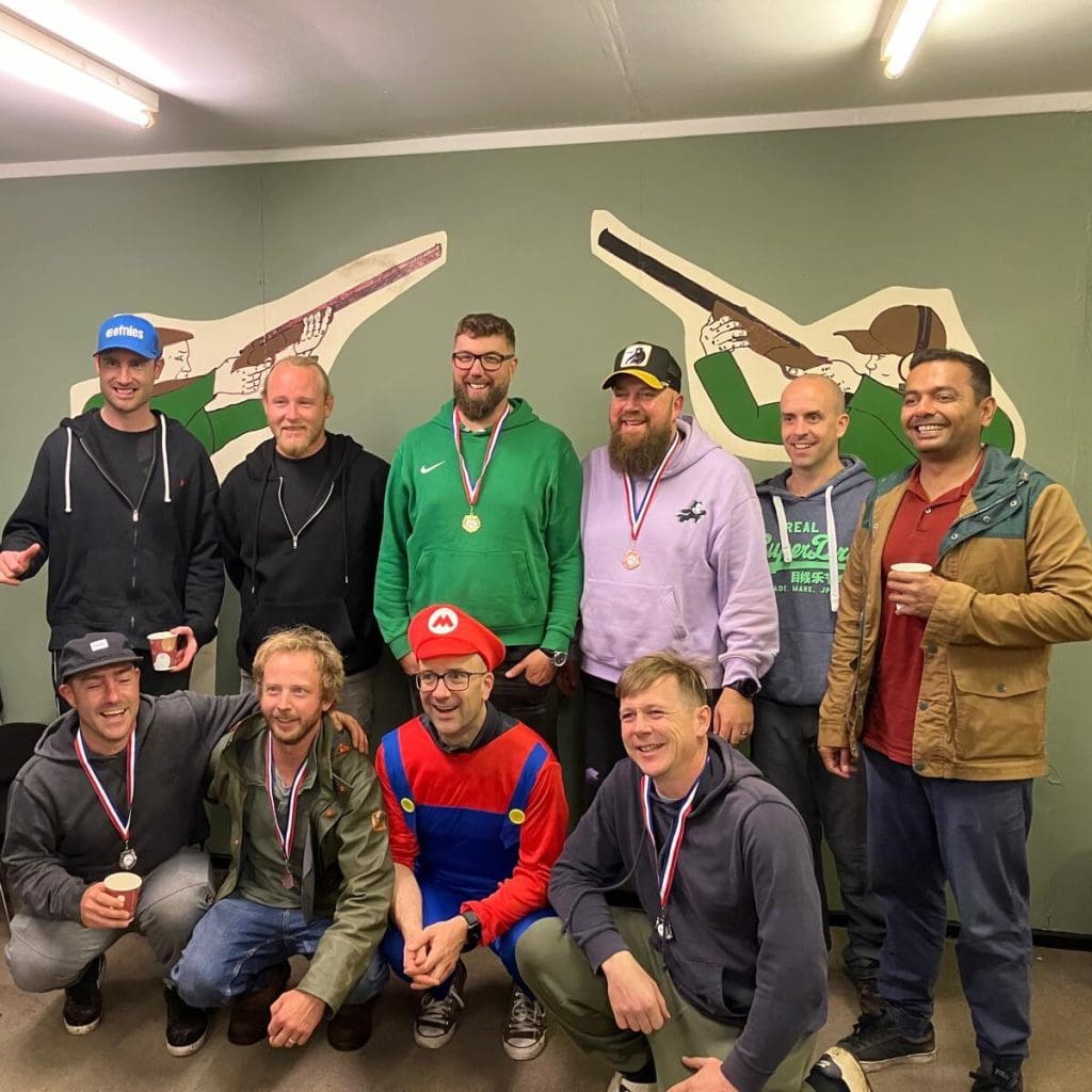 A group of ten smiling men, some wearing medals, pose in front of a green wall with illustrations of shooters. One man is dressed in a Mario costume. Everyone appears happy, and some are holding drinks.