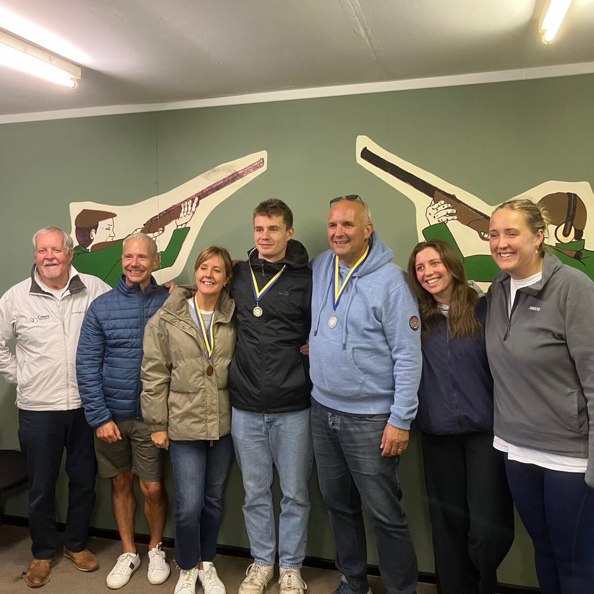 Seven people stand in a row indoors, some wearing medals around their necks. They are smiling in front of a green wall decorated with painted images of people holding rifles.