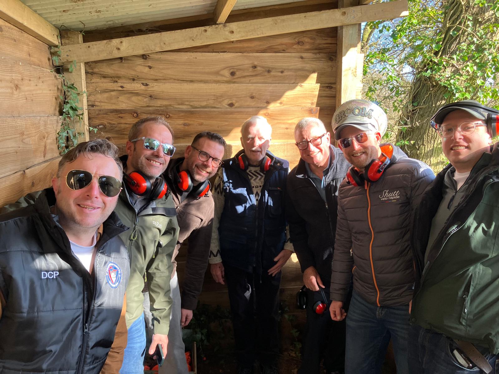 Seven men wearing jackets and sunglasses, some with headphones around their necks, stand closely together inside a wooden shelter with sunlight shining in, smiling at the camera.