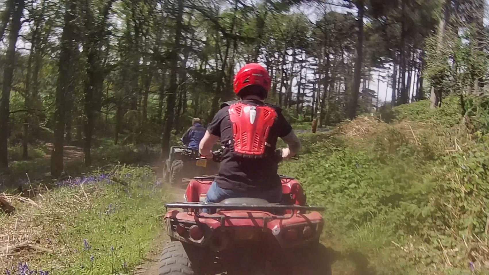 A person wearing a red helmet and red protective gear rides an ATV on a dirt trail through a forest, following another rider ahead. Sunlight filters through the trees and dust rises from the ground.